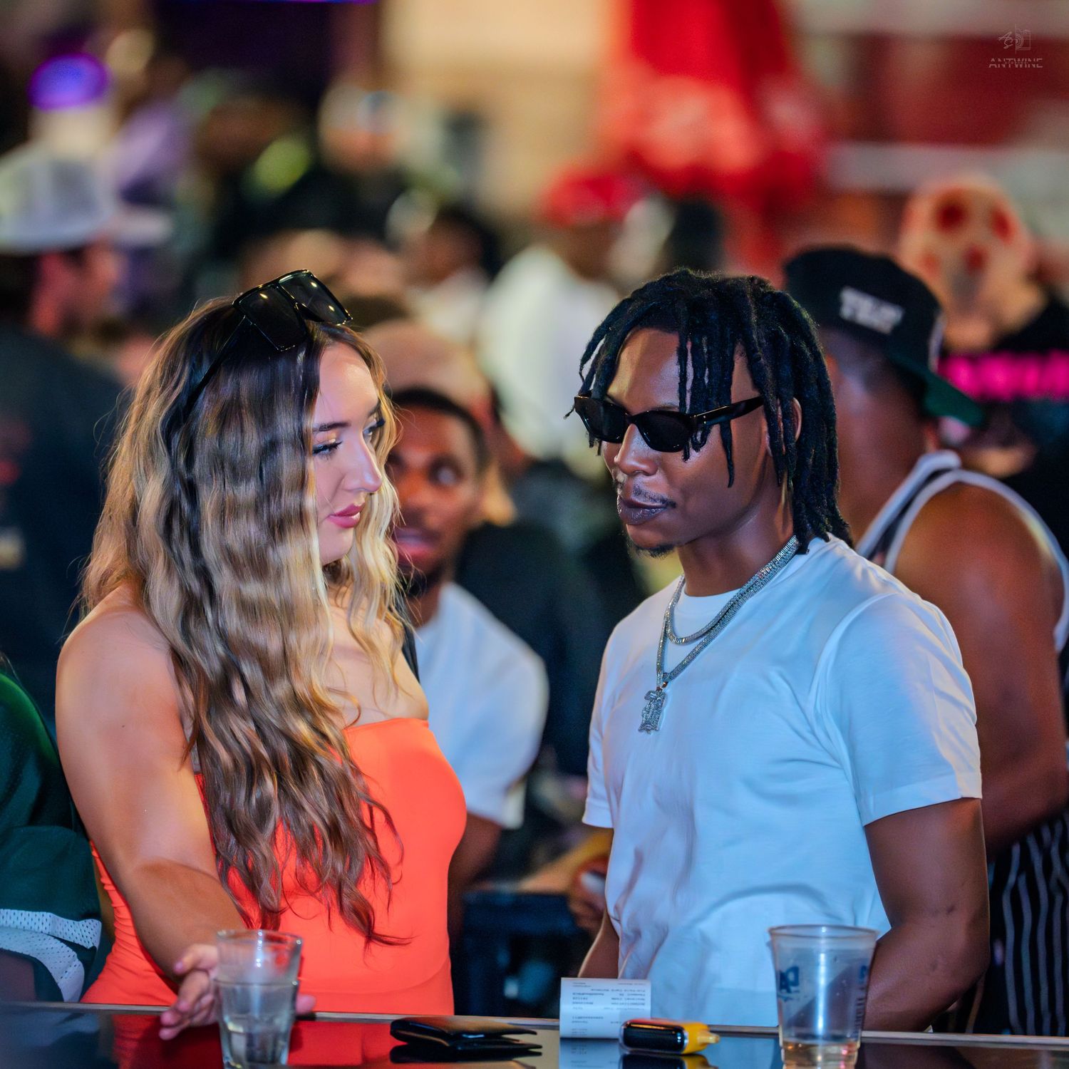 A woman in an orange top and a man in a white shirt chat at a crowded nightclub event.