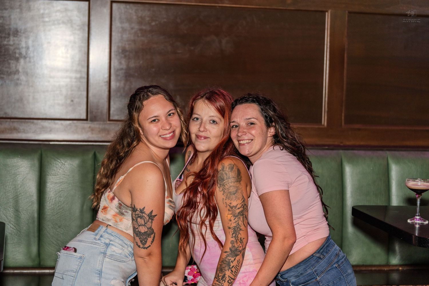 Three tattooed women smiling and posing together in a bar booth with green seating.
