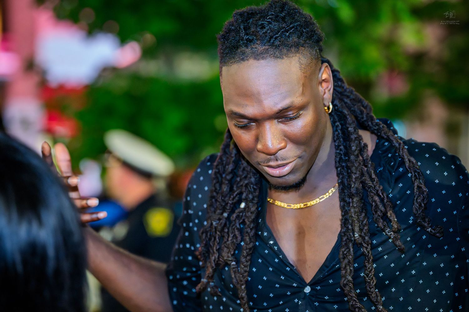 A tall Black man with dreadlocks wearing a black polka dot shirt and gold necklace looks down at an outdoor event.