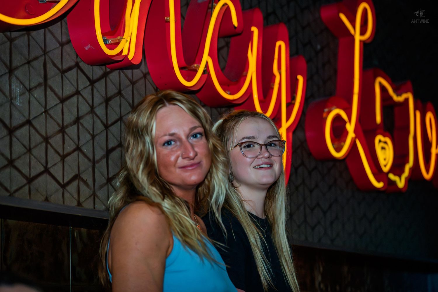 Two blonde women posing together in front of a glowing neon sign at a bar.