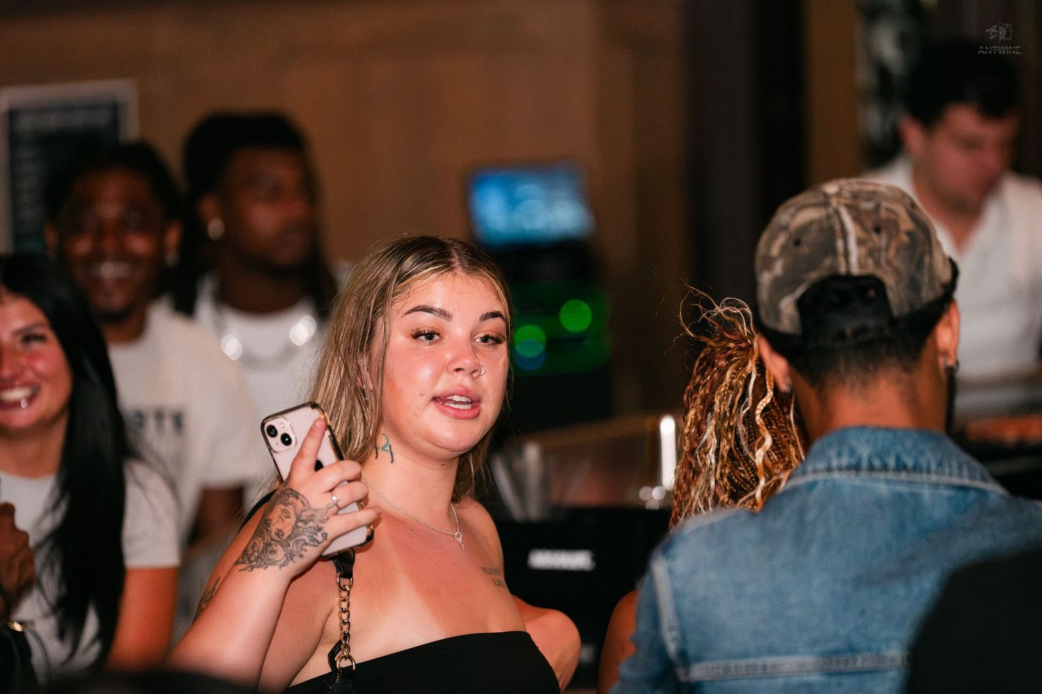 Blonde woman holding a pink phone at a lively indoor party event.