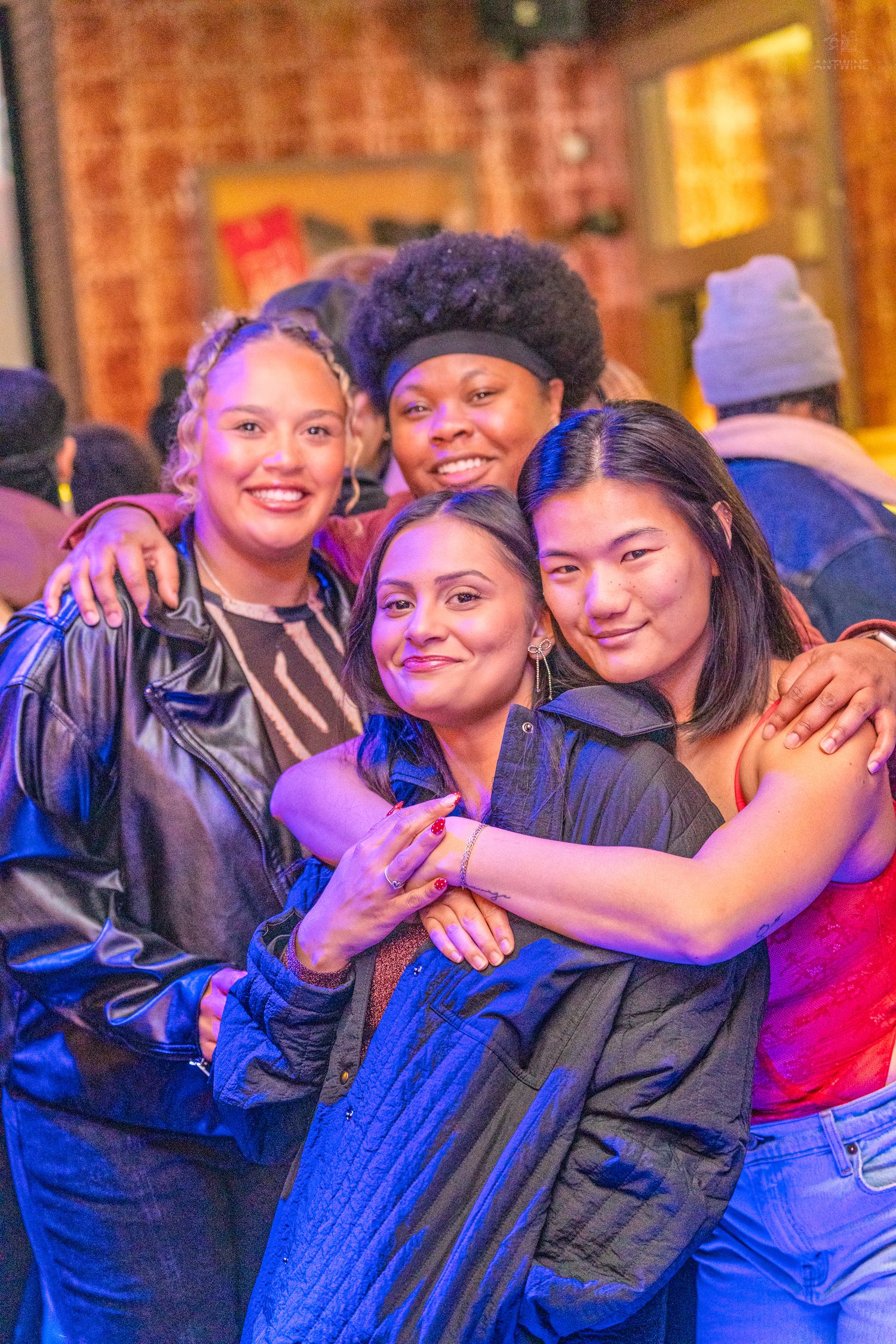 Four smiling women embrace each other under purple lights at an outdoor nighttime gathering.