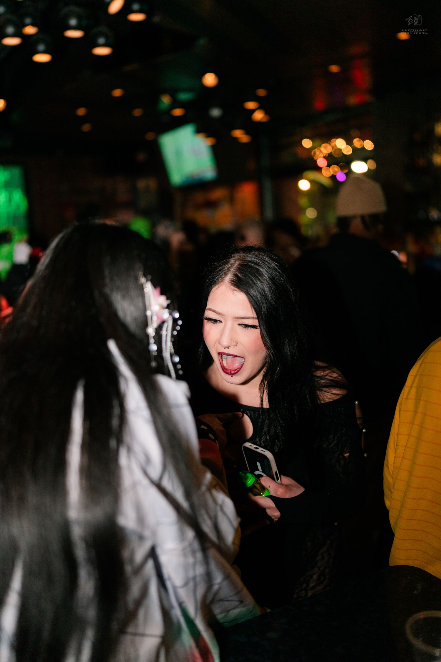 Young woman in black Halloween costume sticking out tongue while holding phone at a nightclub party.