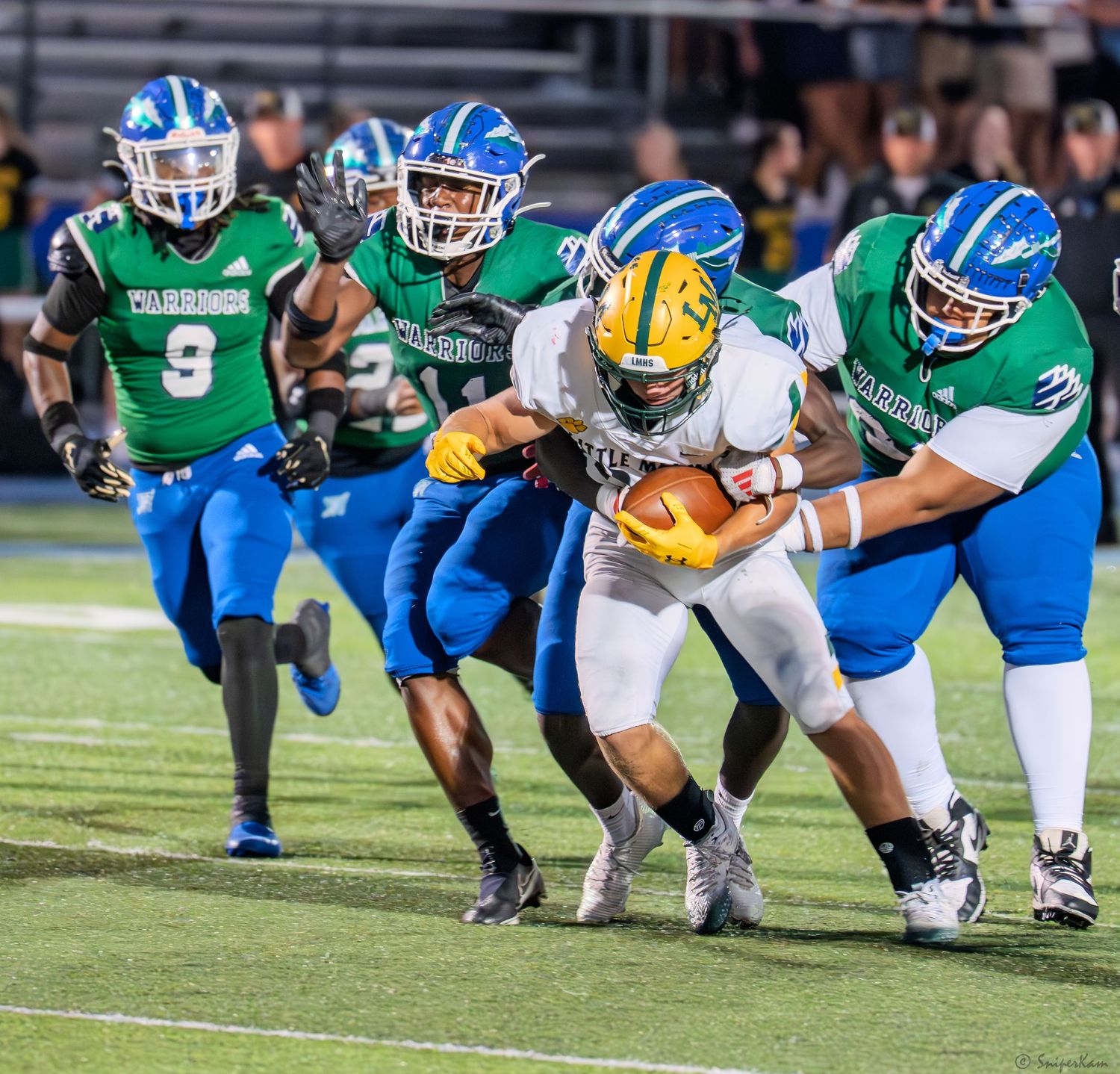 Multiple Warriors defenders in green jerseys tackle a yellow-uniformed ball carrier during a football game.
