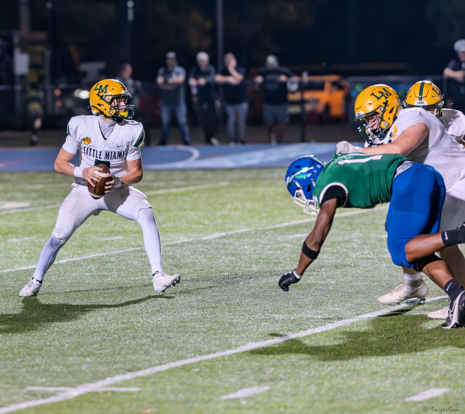 A quarterback in white and gold uniform scans the field while an offensive lineman blocks a green-uniformed defender.