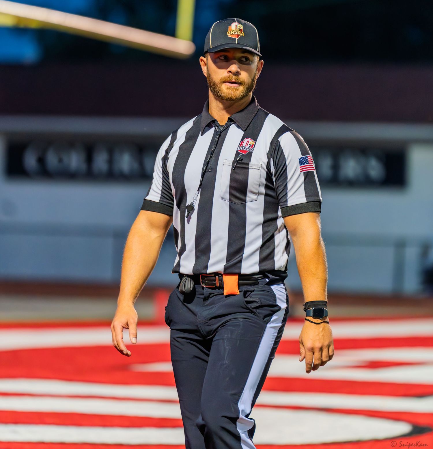 High school football referee in black and white striped uniform walks the field during a night game.