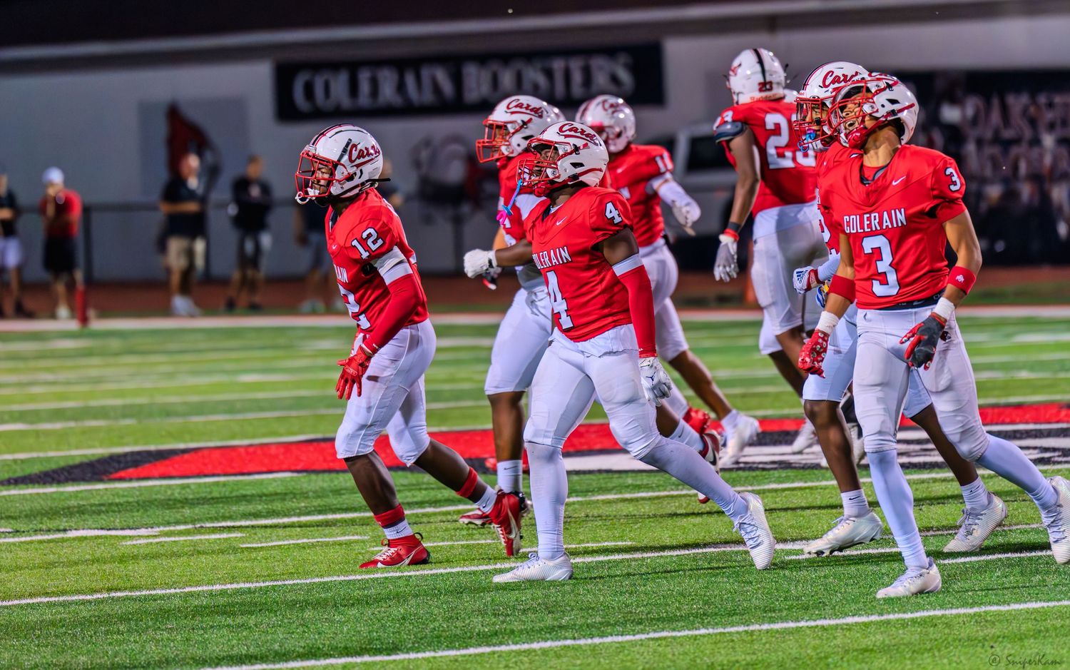 Colerain high school football players in red uniforms celebrate on the field during a night game.