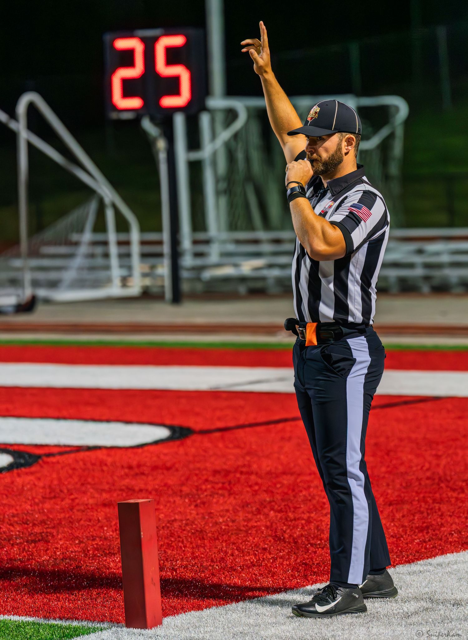 Football referee signals with one finger raised near a red pylon during a night game with score 25.