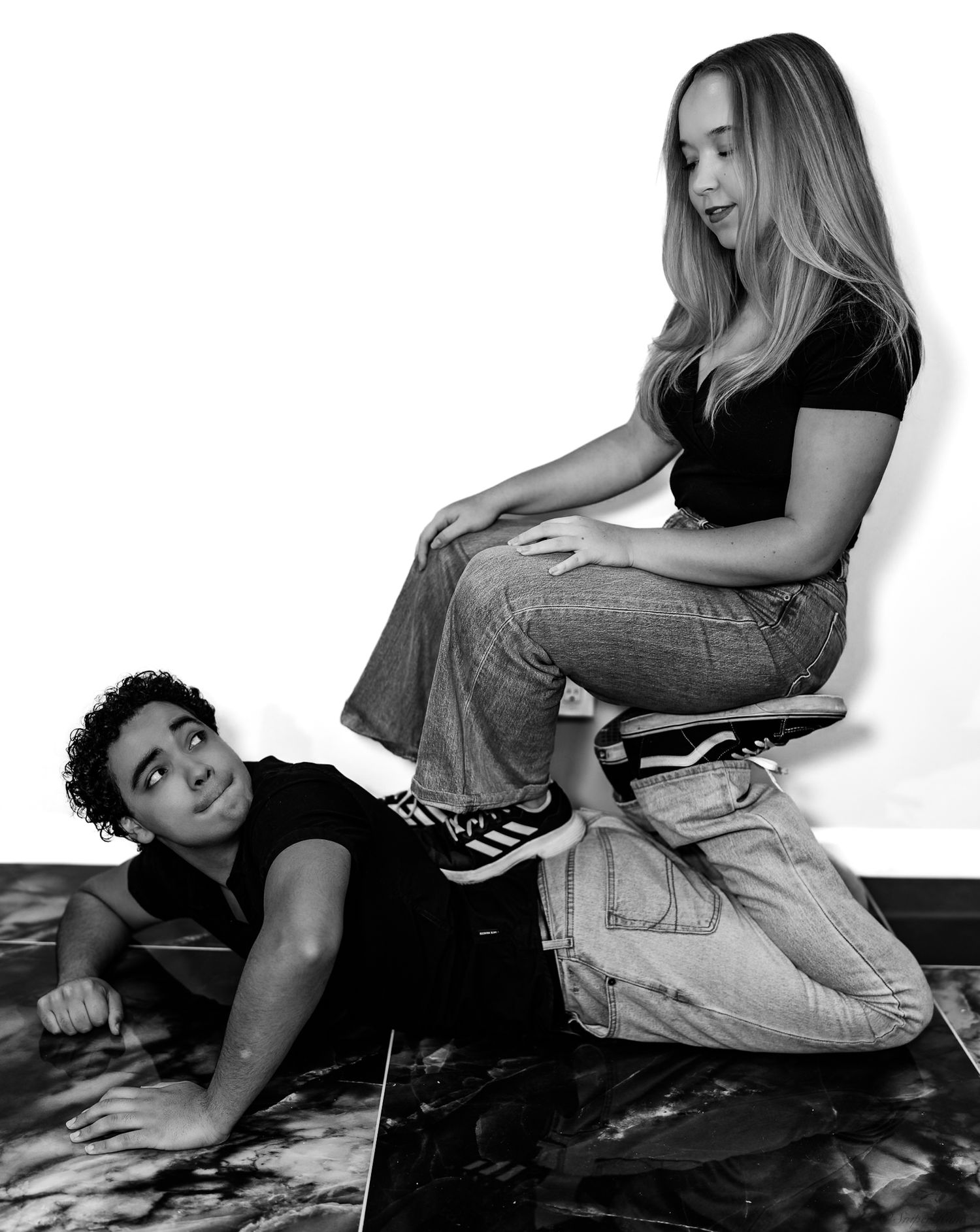 Black and white photo of a young woman sitting on a stool while a young man lies on the floor beside her.