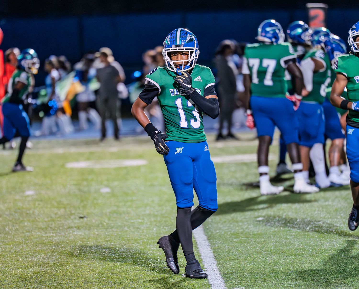 Football player number 13 in green and blue uniform stands on sideline during night game.