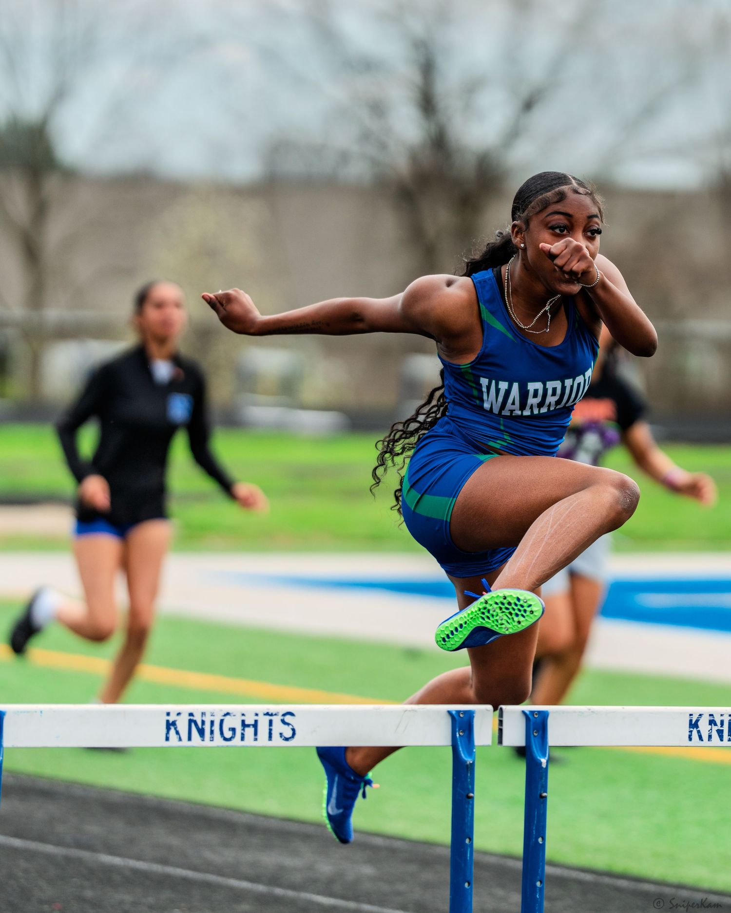 Female Warriors athlete clearing a hurdle during a track and field competition at Knights stadium.