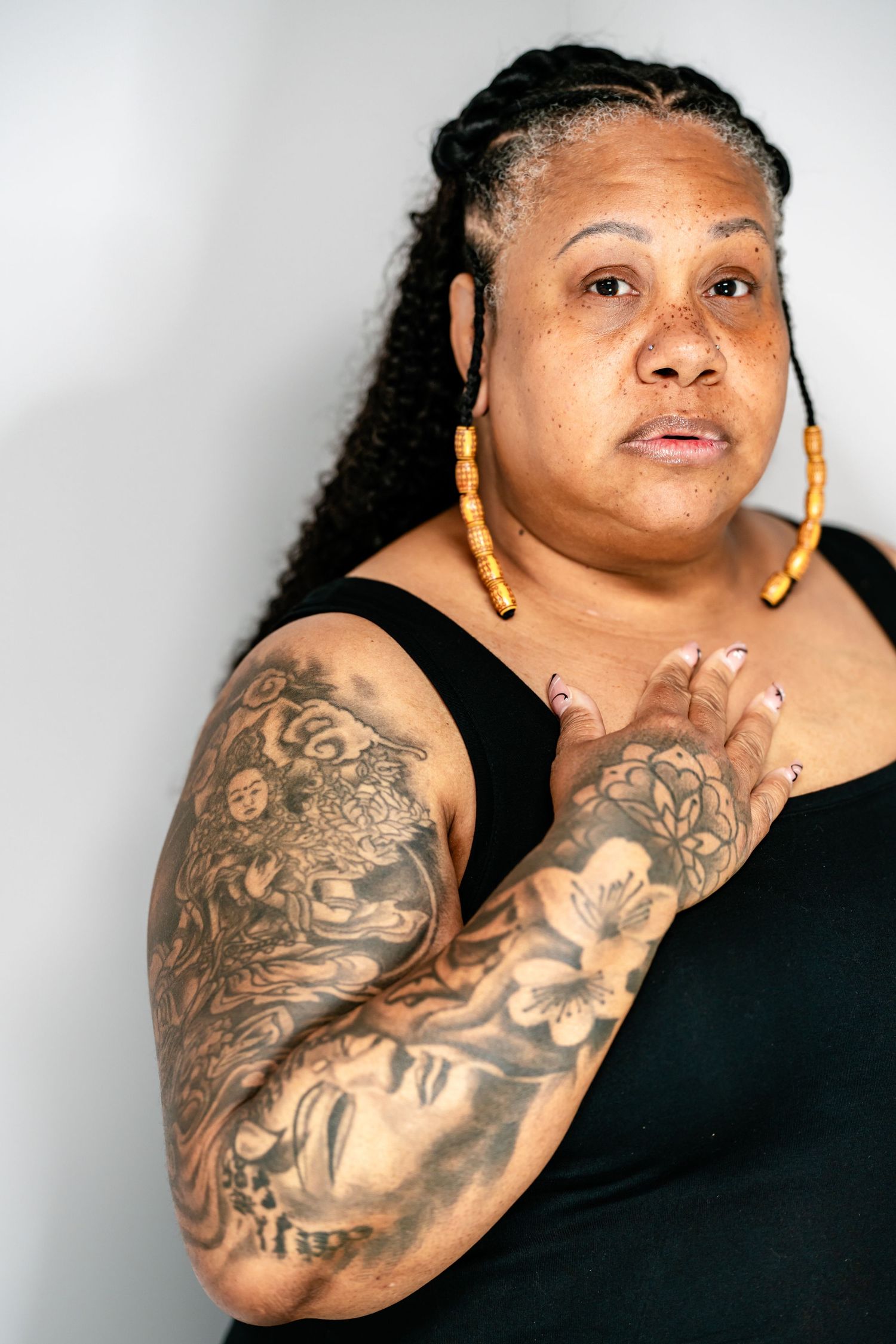A Black woman with braided hair and gold earrings displays detailed sleeve tattoos in a color studio portrait.