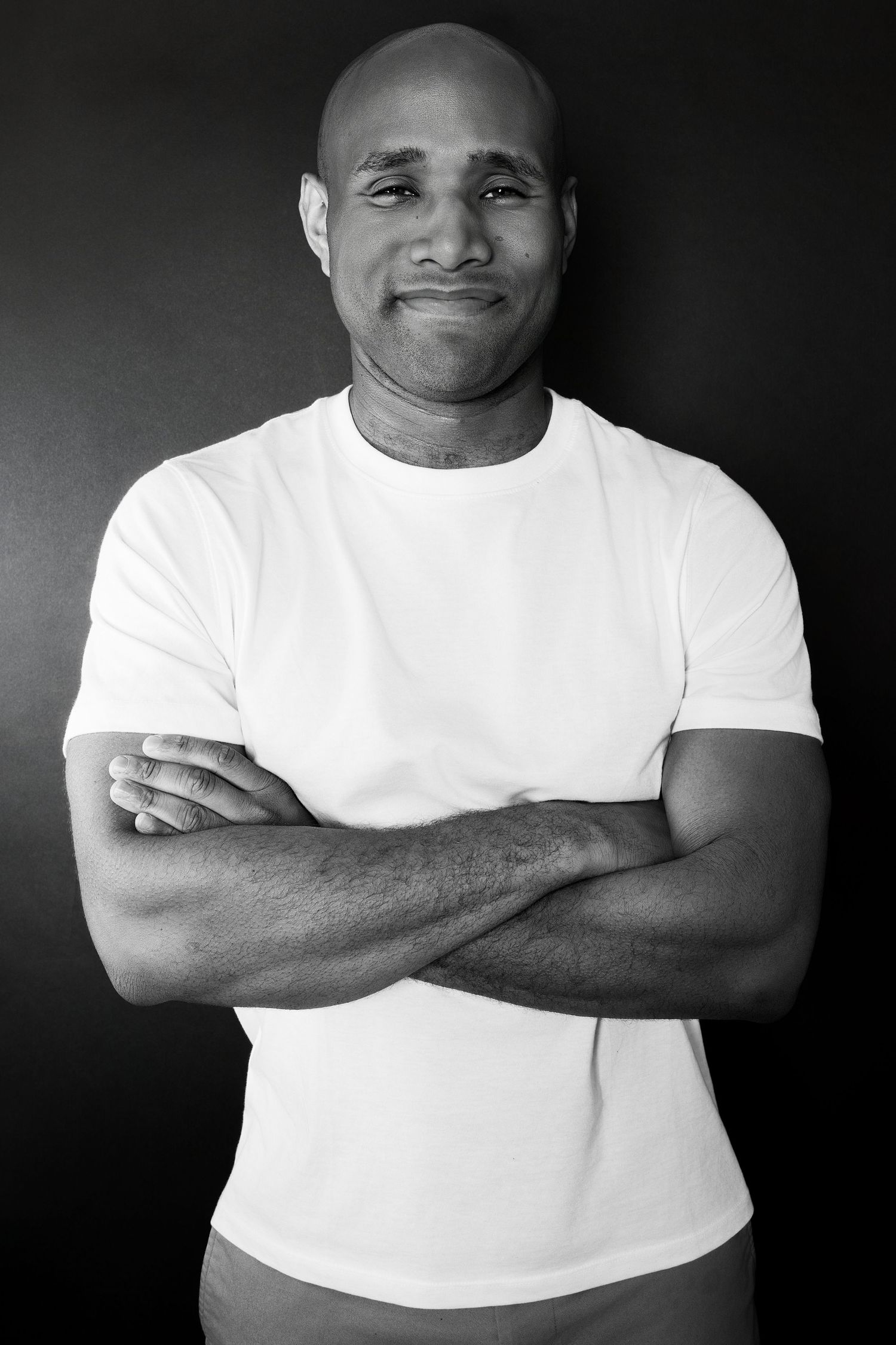 Muscular bald Black man in white t-shirt standing with arms crossed in black and white photo.