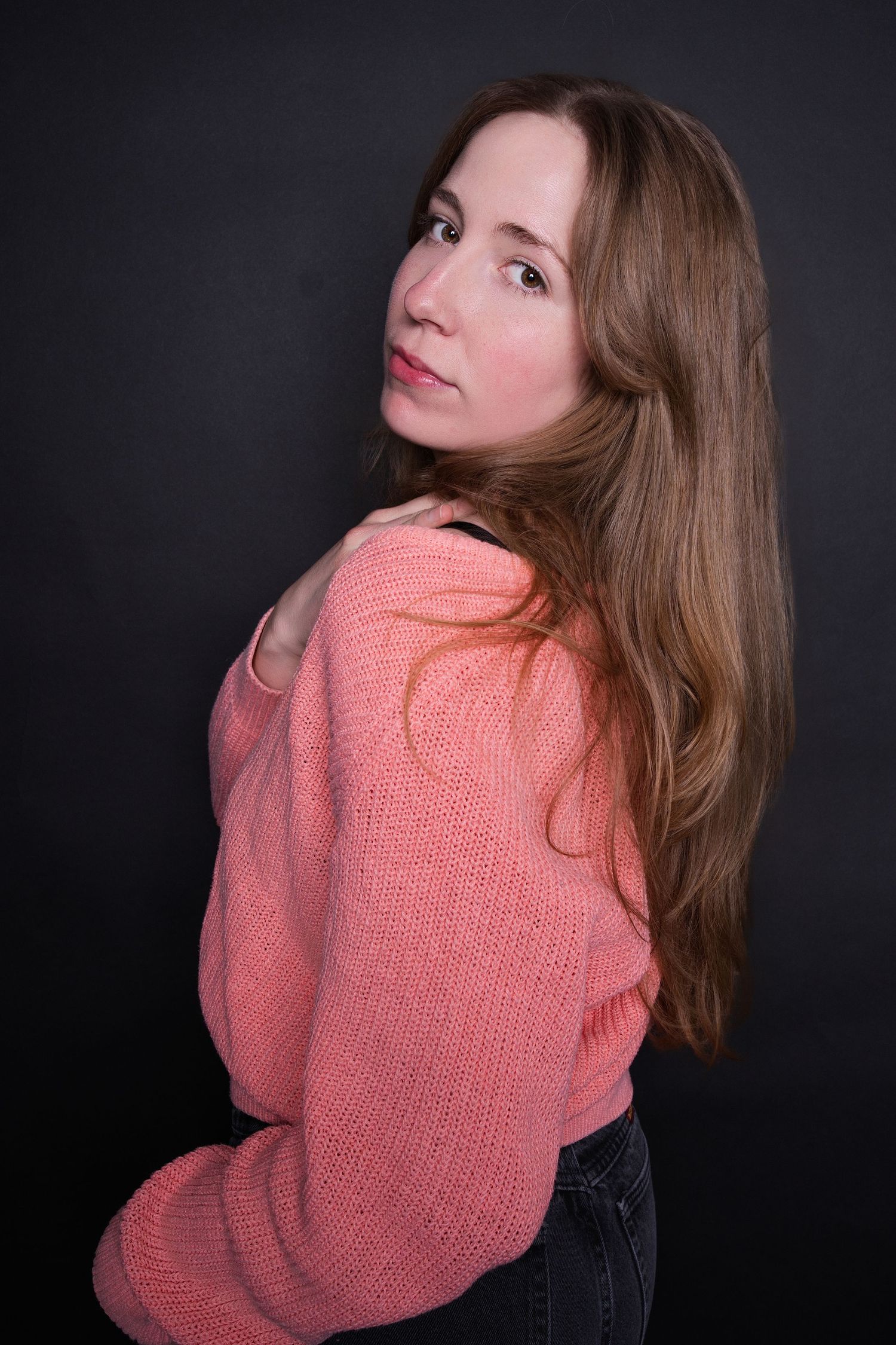 Woman with long brown hair in a pink knit sweater looking over her shoulder against a dark background.