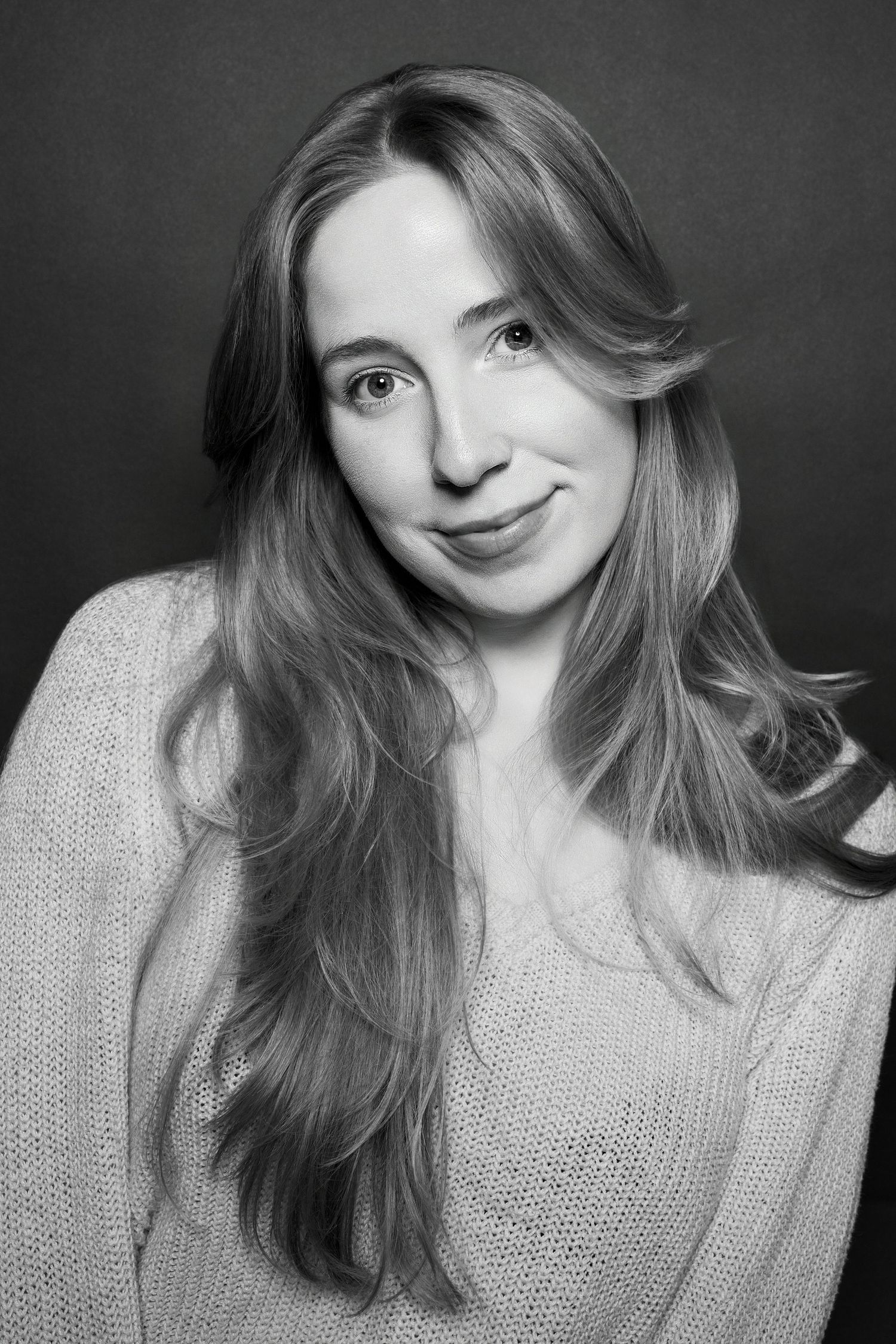 Black and white studio portrait of a smiling young woman with long hair in a cozy knit sweater.