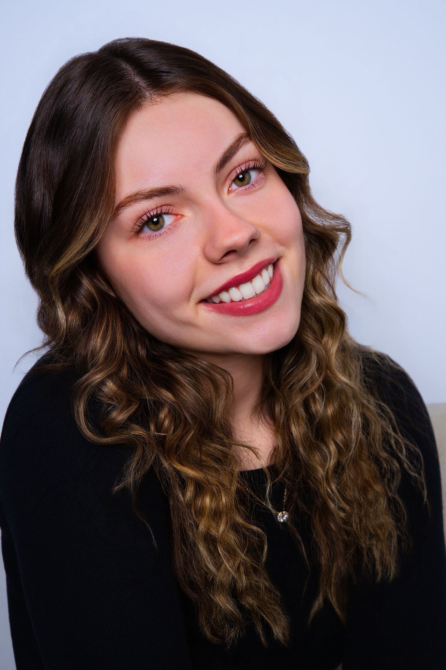 Smiling young woman with curly brown highlighted hair wearing a black top and dainty necklace.