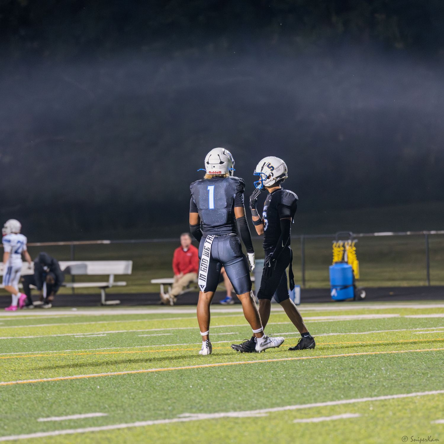 Two football players in dark uniforms converse on a lit field during a night game.