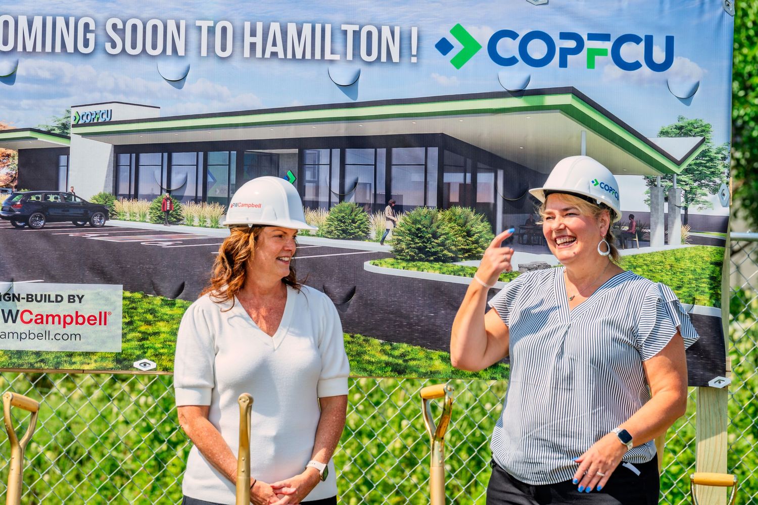Two women in hard hats smile in front of COPFCU Coming Soon to Hamilton construction rendering sign.
