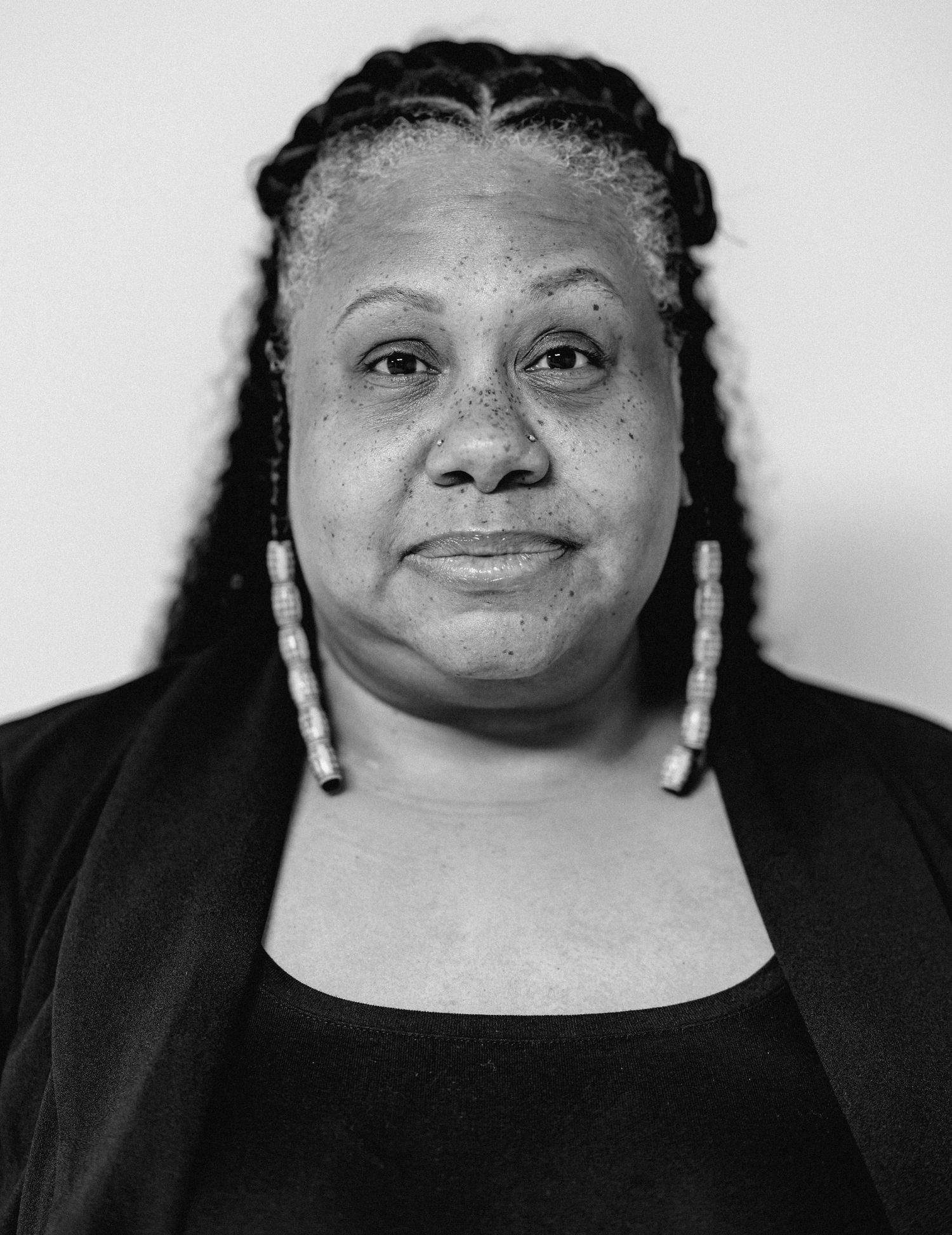 Black and white portrait of a smiling woman with long braids and dangling earrings in black top.