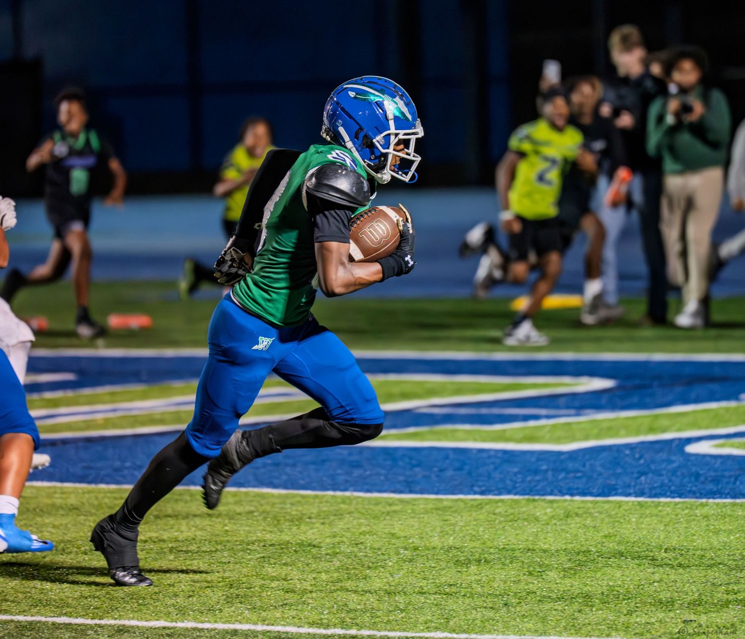 Football player in green and blue uniform runs with the ball on a blue turf field during a night game.