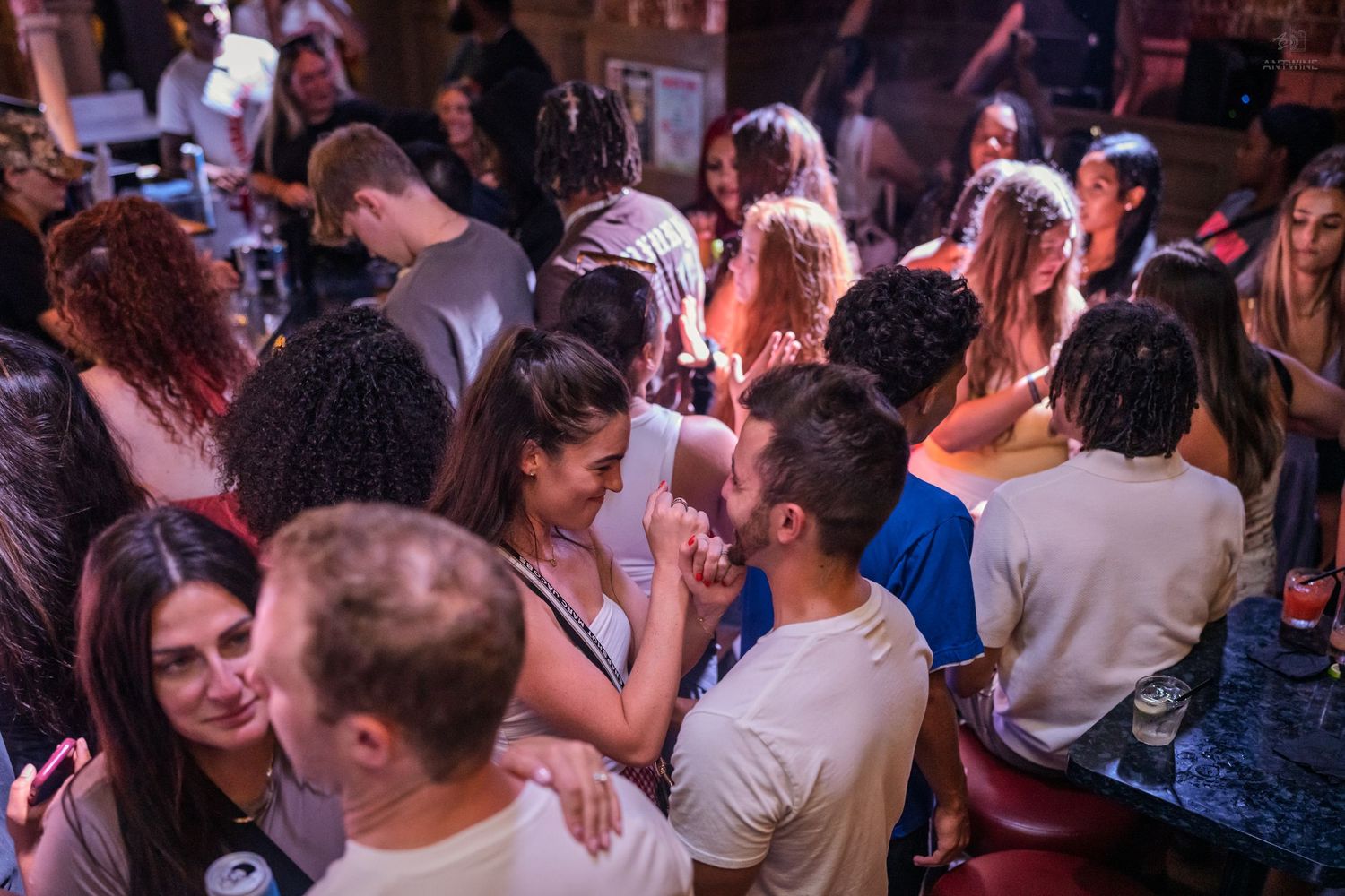 Busy nightclub bar scene with groups of young adults socializing under warm pink and red lighting.