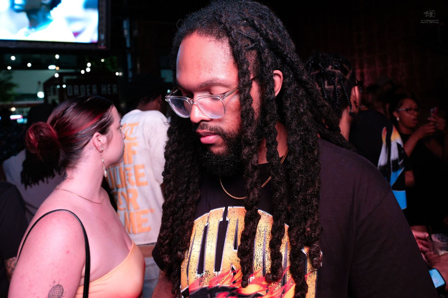 Man with long dreadlocks and glasses standing at a nightclub event wearing a graphic tee.