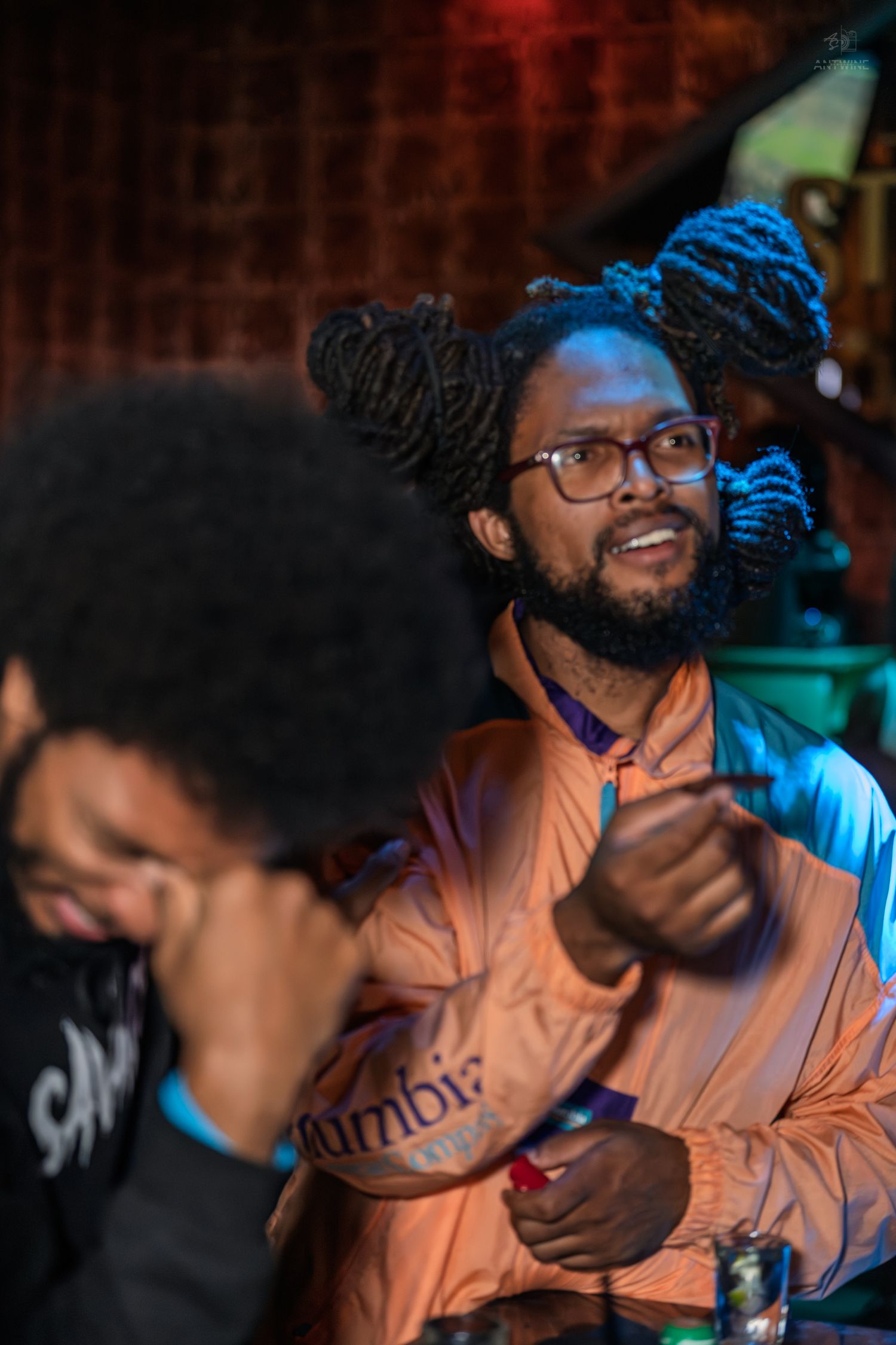 Smiling man with dreadlocks and glasses wearing a Columbia jacket at a lively bar event.