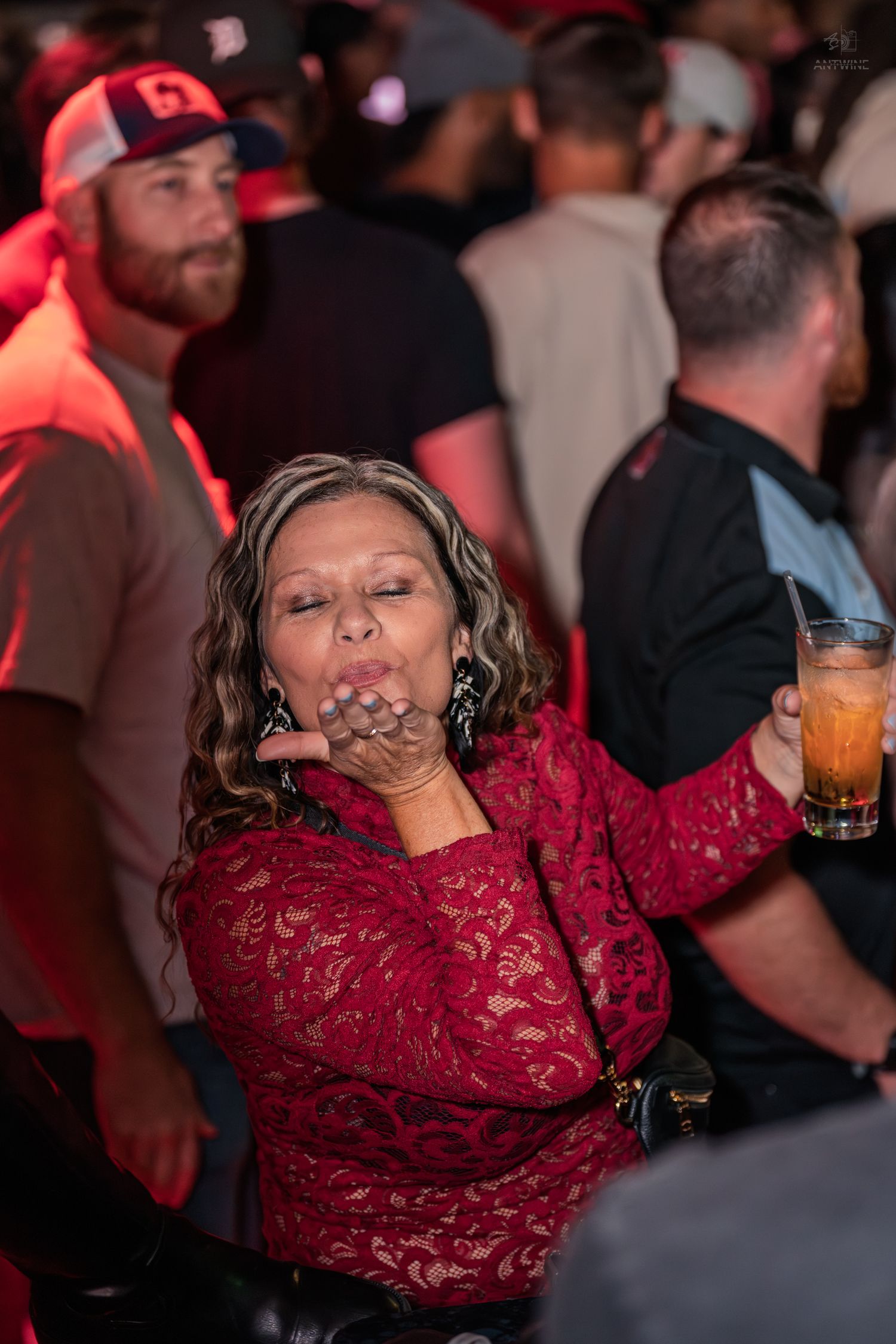 Woman in red lace dress blowing a kiss while holding a drink at a busy nightclub.