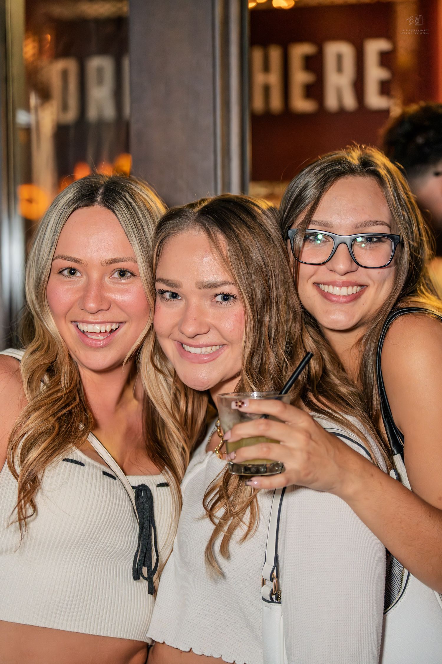 Three smiling women in white tops pose together outside a bar holding drinks.