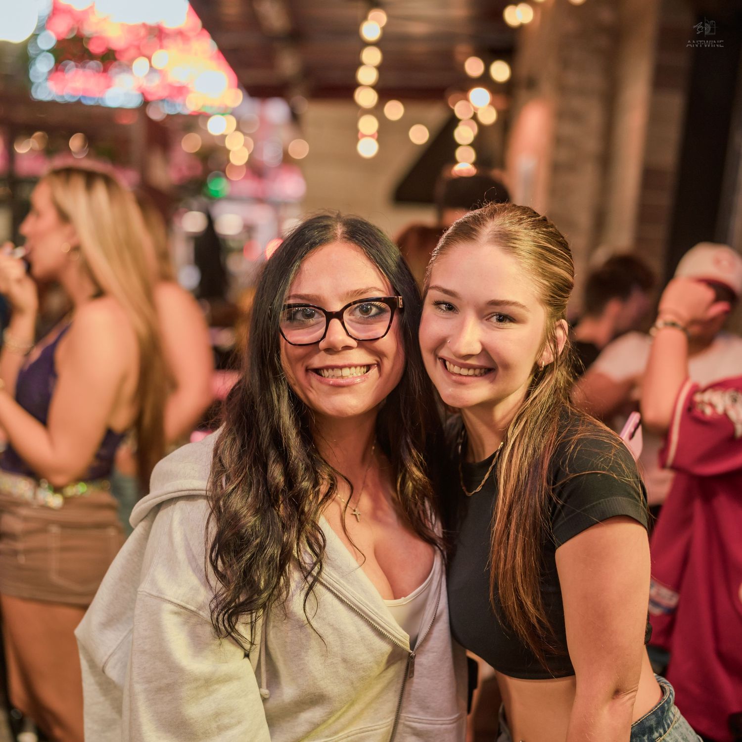 Two women smile and pose together on a bar patio at night with string lights in the background.