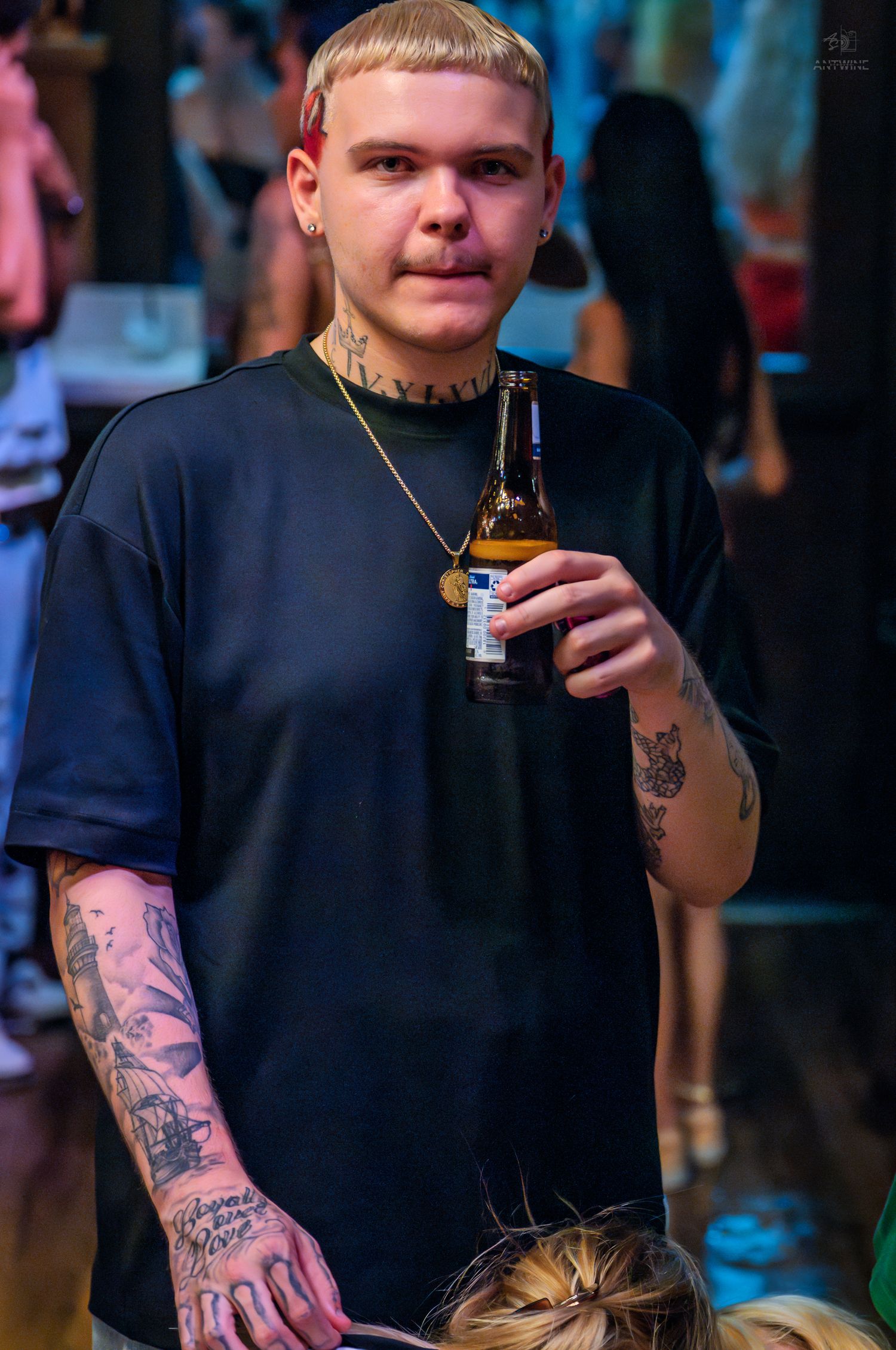 Tattooed young man with blonde hair holding a beer bottle at a nightclub event.