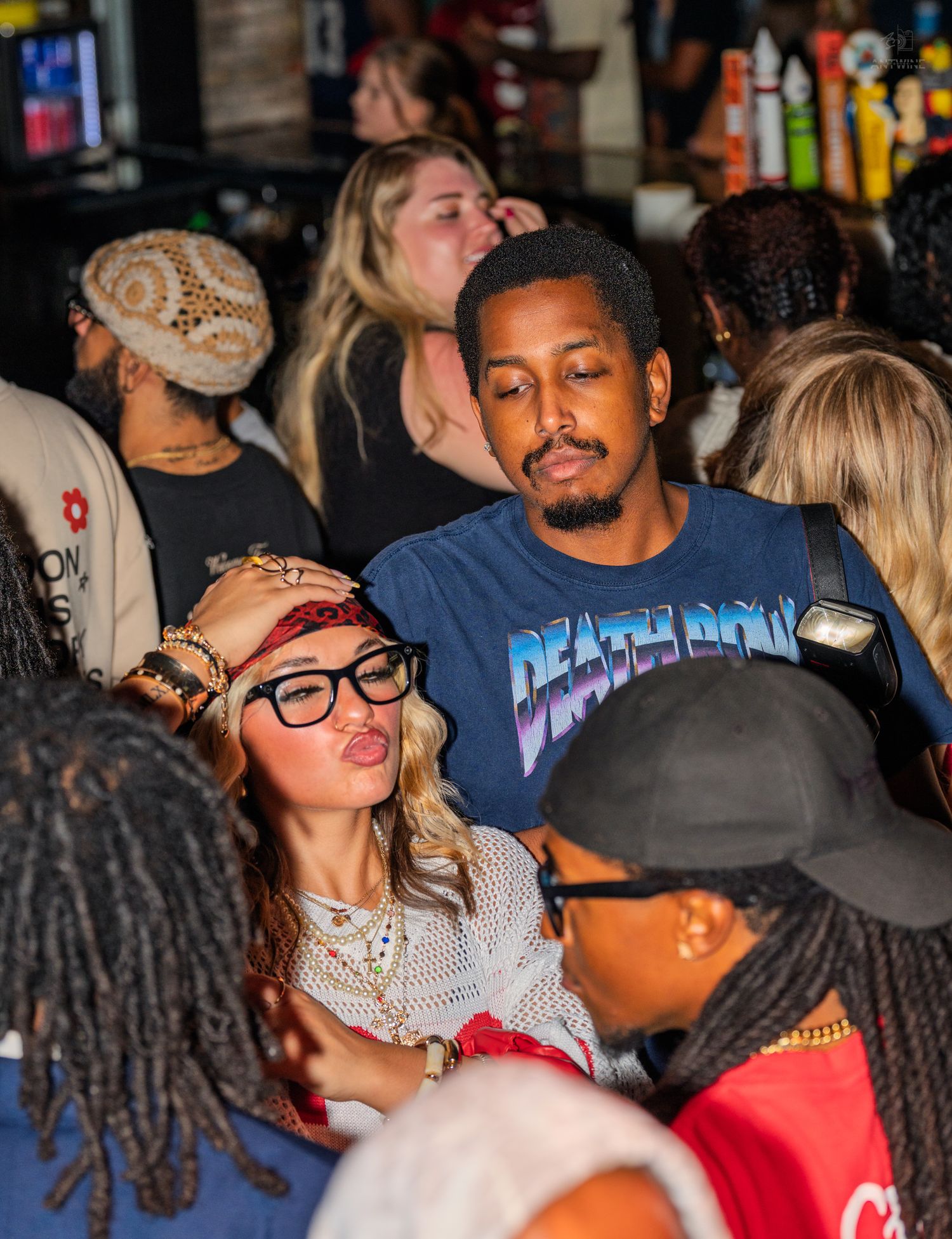Man in Death Row Records shirt socializing with woman in glasses at a crowded bar.