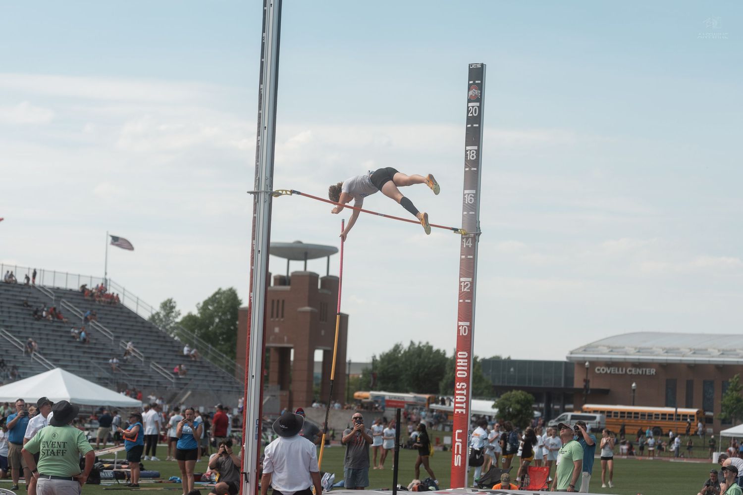 Athlete performs a pole vault at an outdoor track and field meet with stadium seating in background.