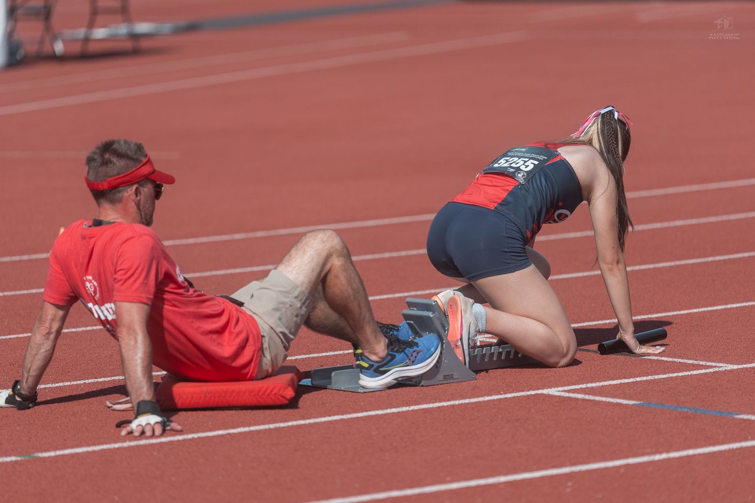 Female sprinter in starting position on red track with male coach lying beside starting blocks.