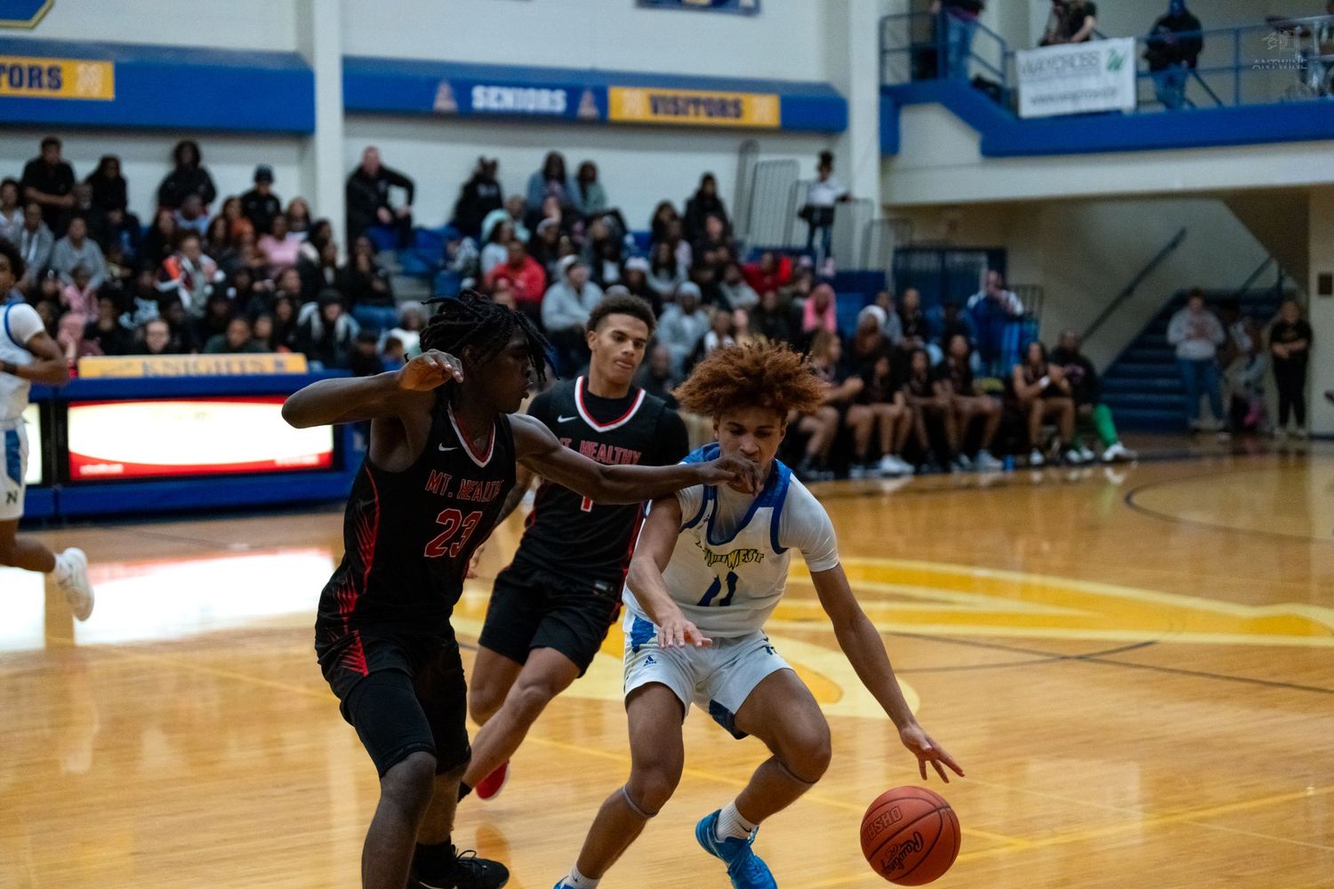 High school basketball player number 11 in blue drives past two defenders in red during an indoor game.