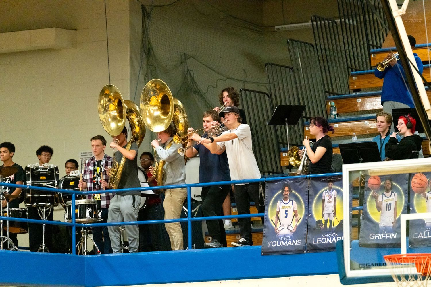 High school pep band plays tubas and brass instruments on a blue balcony during a basketball game.