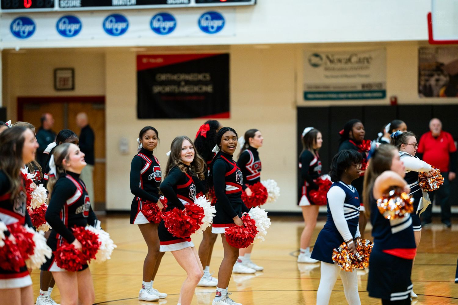High school cheerleaders in red and black uniforms smile while performing with pom-poms in a gymnasium.