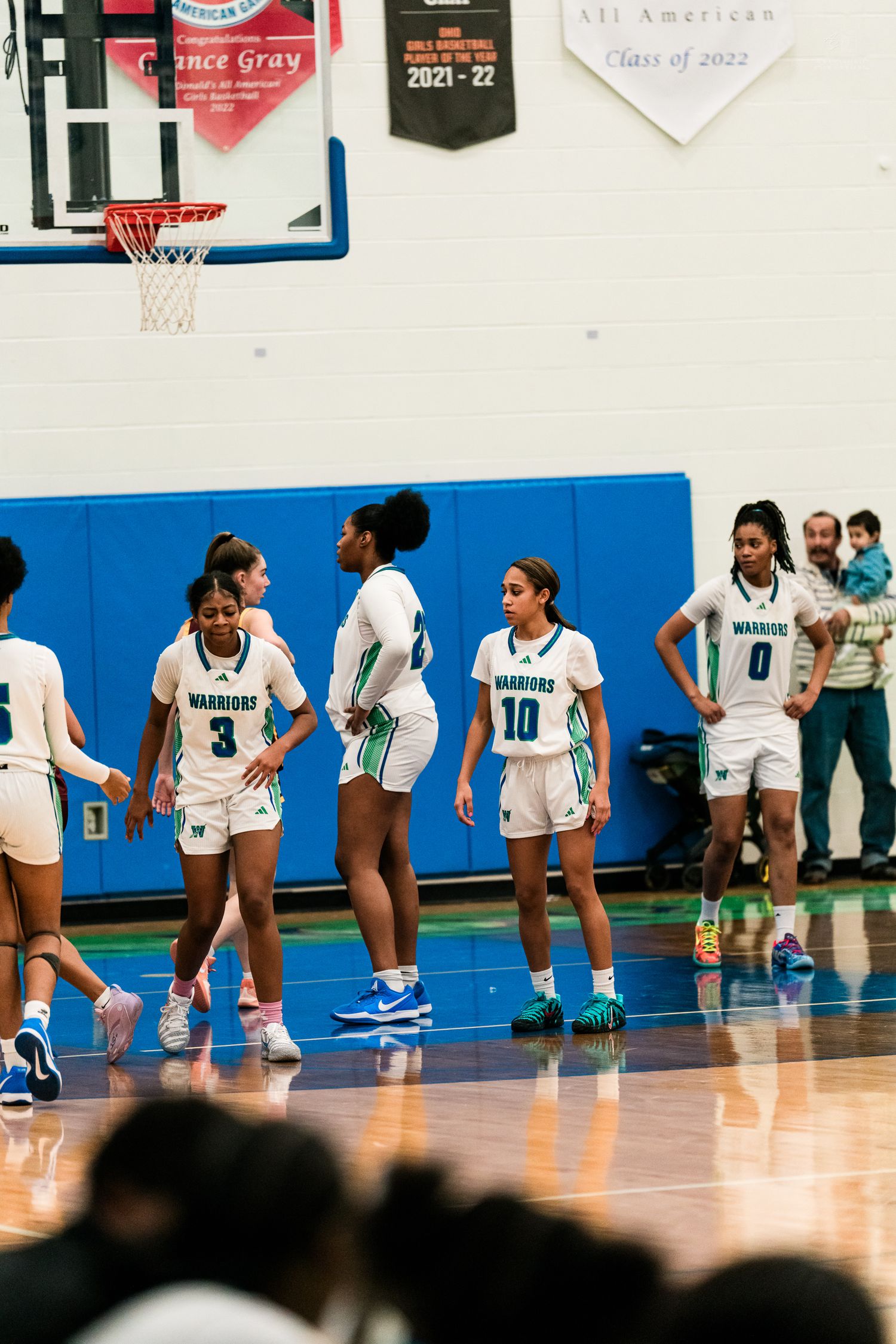 Warriors girls basketball team huddles on court in white and teal uniforms during a high school game.