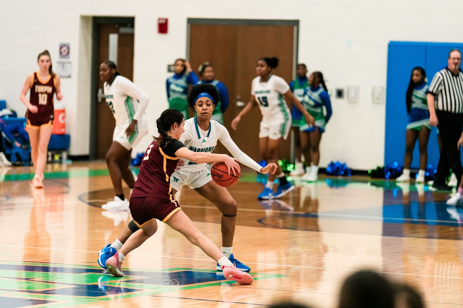Warriors defender pressures Trojan ball handler during girls high school basketball game with cheerleaders in background.