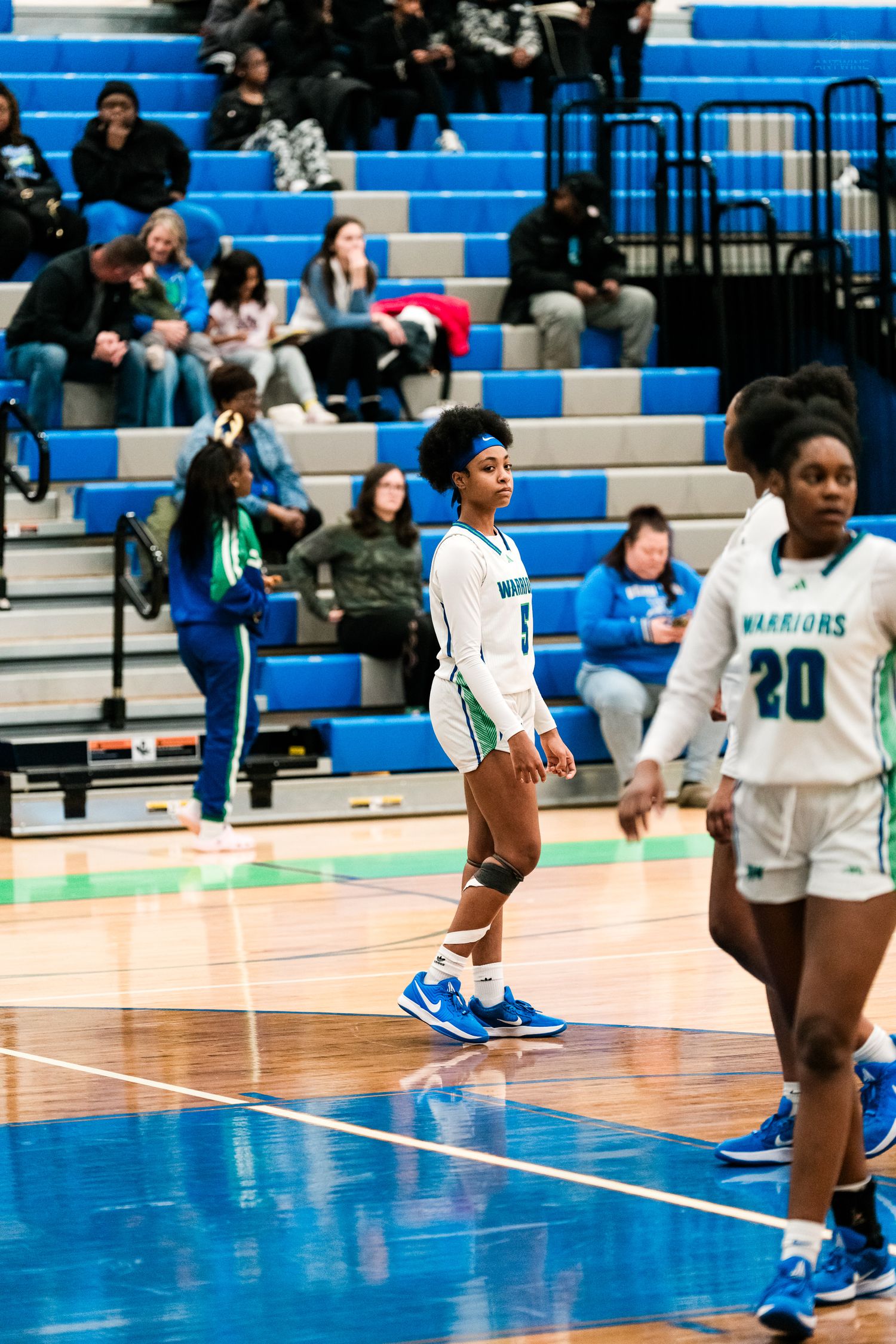 Warriors female basketball player walks on hardwood court in white teal uniform during high school game.