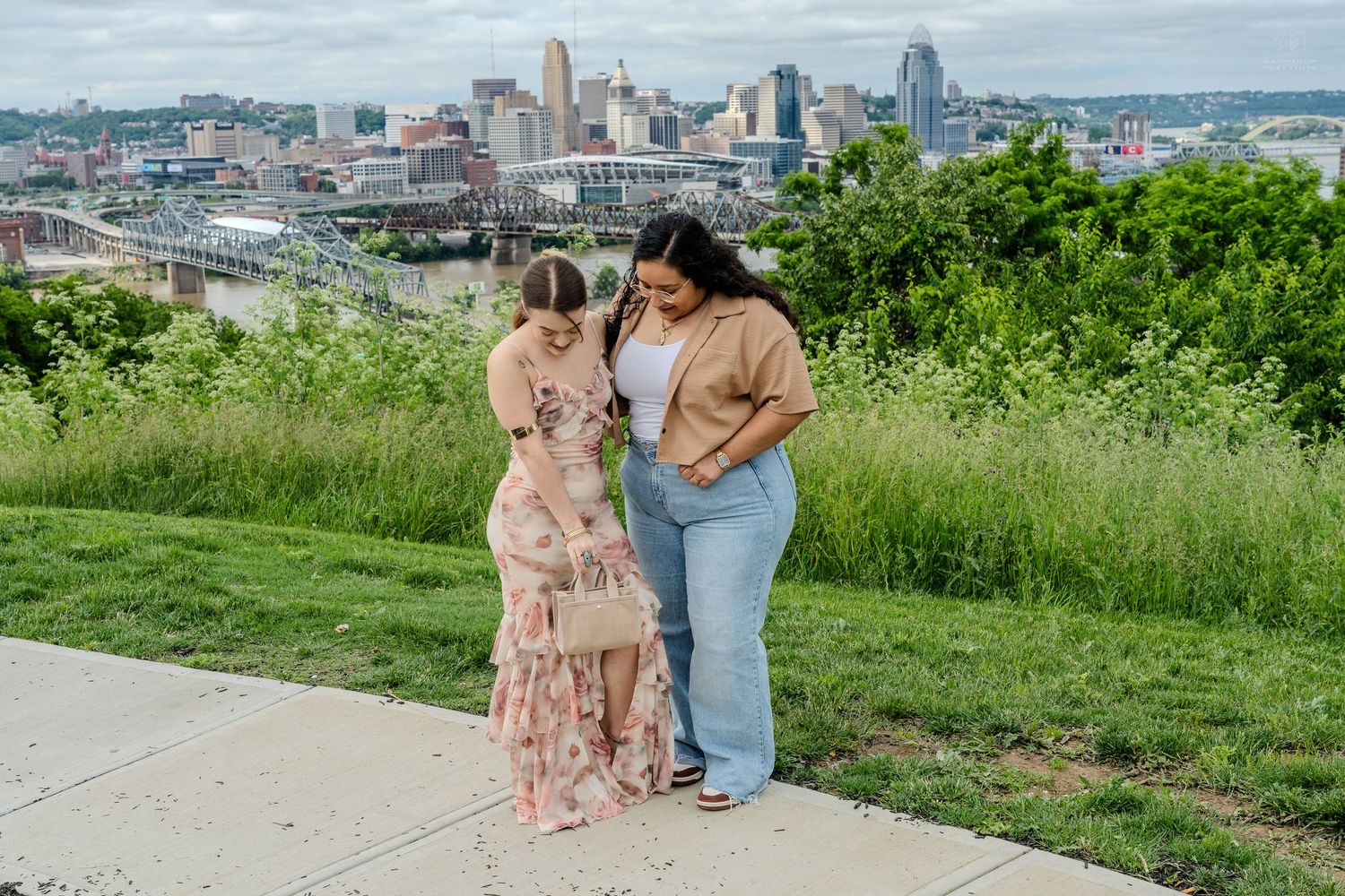Two women laughing together outdoors with colorful Cincinnati city skyline in background.