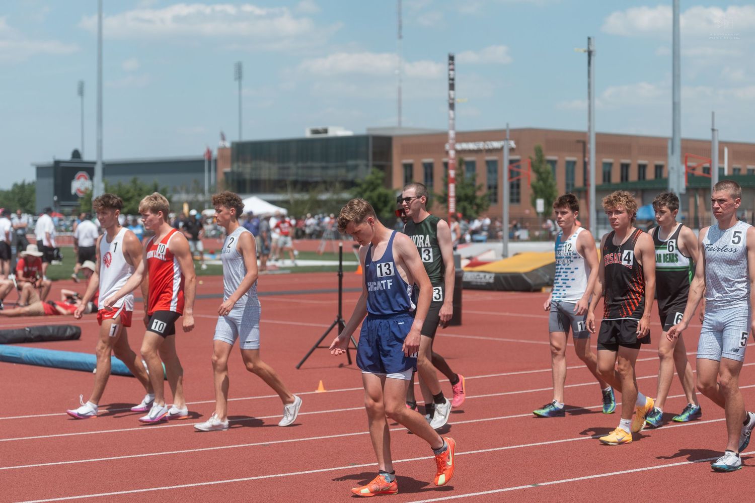 Male track athletes in various uniforms walk along a red track at an outdoor field meet.