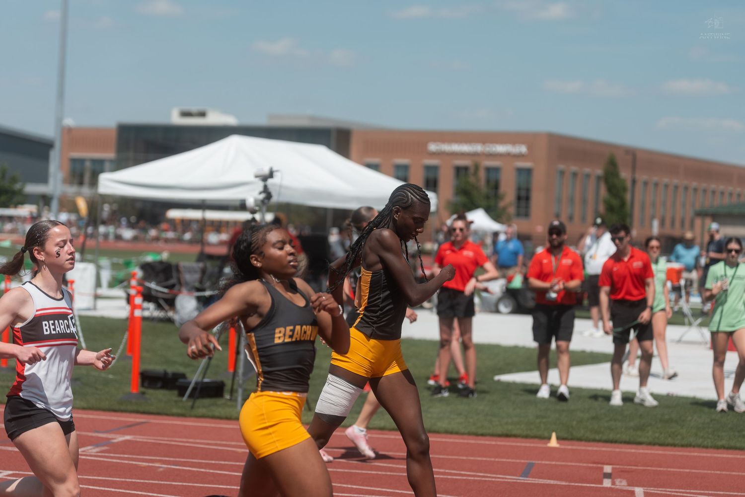 Two female athletes in gold and black uniforms perform a baton exchange during a high school relay race.