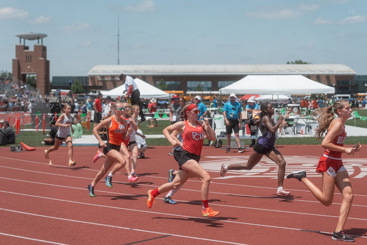 Young women runners race on a red track at an outdoor athletic competition with spectators.