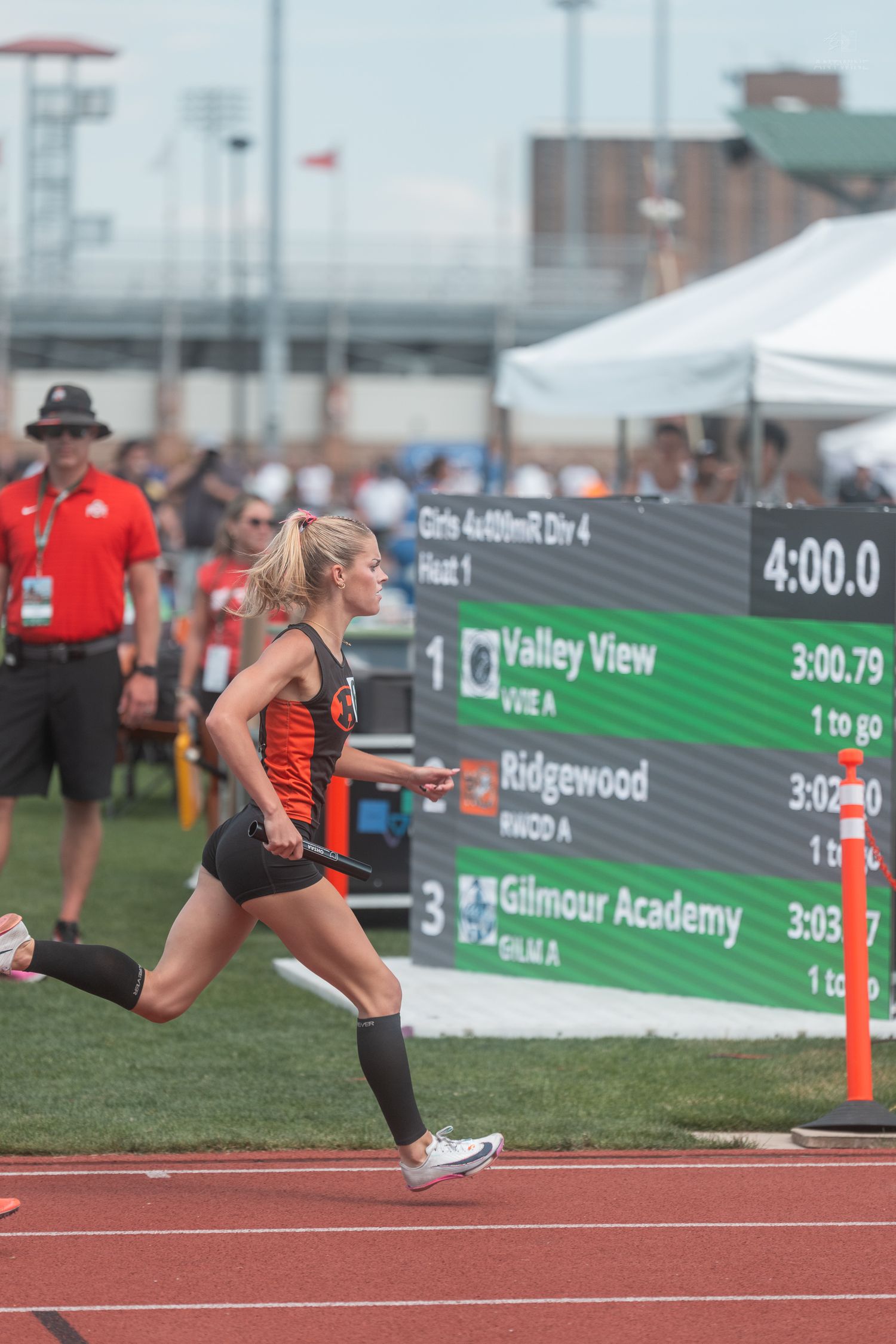 Female relay runner in orange and black uniform sprints past scoreboard at state track championship.