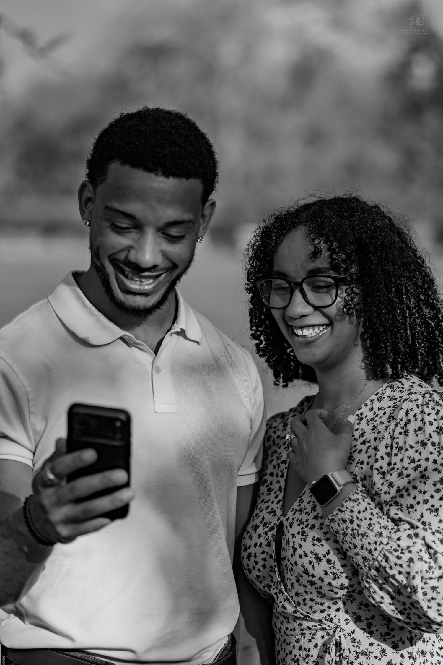 Black and white portrait of laughing couple looking at phone together outdoors in natural setting.