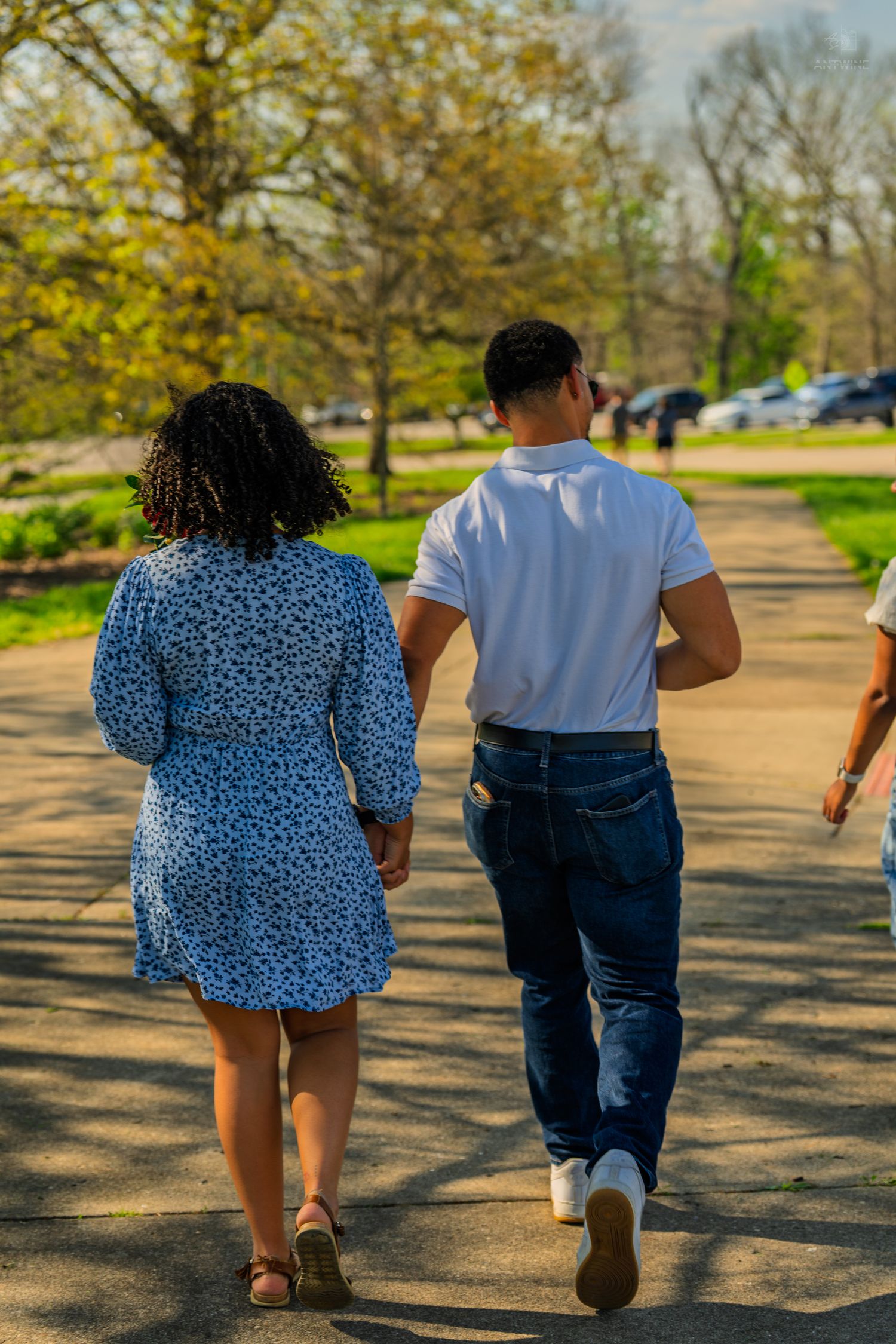 Couple walking hand in hand on a sunlit park path surrounded by green trees in spring.