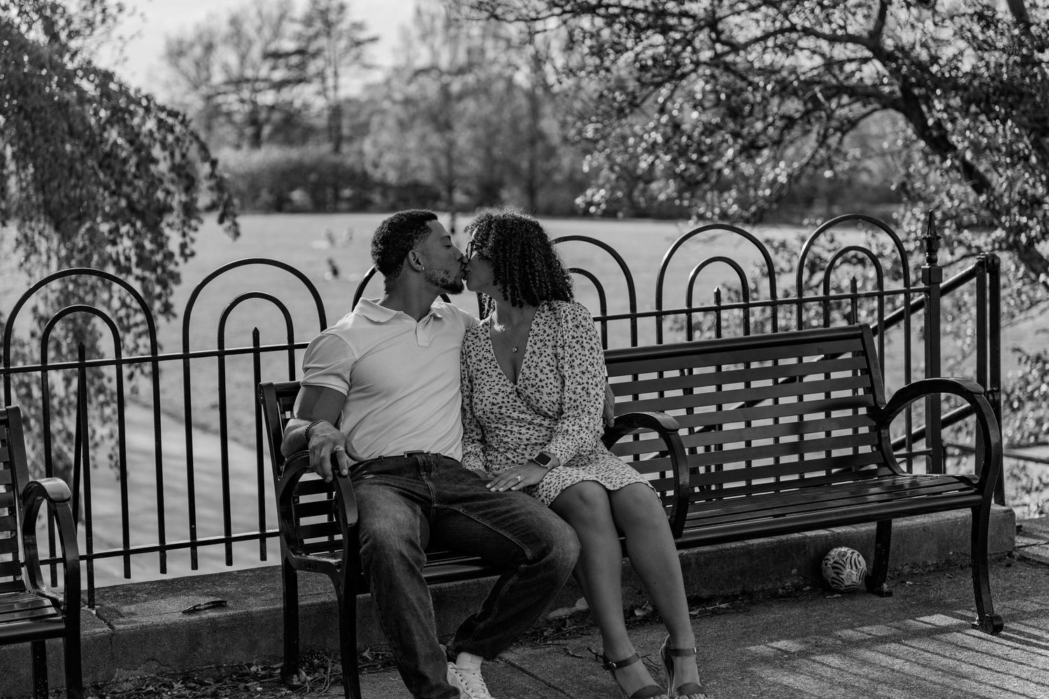 Black and white photo of a couple sharing a kiss on a park bench by a lake.