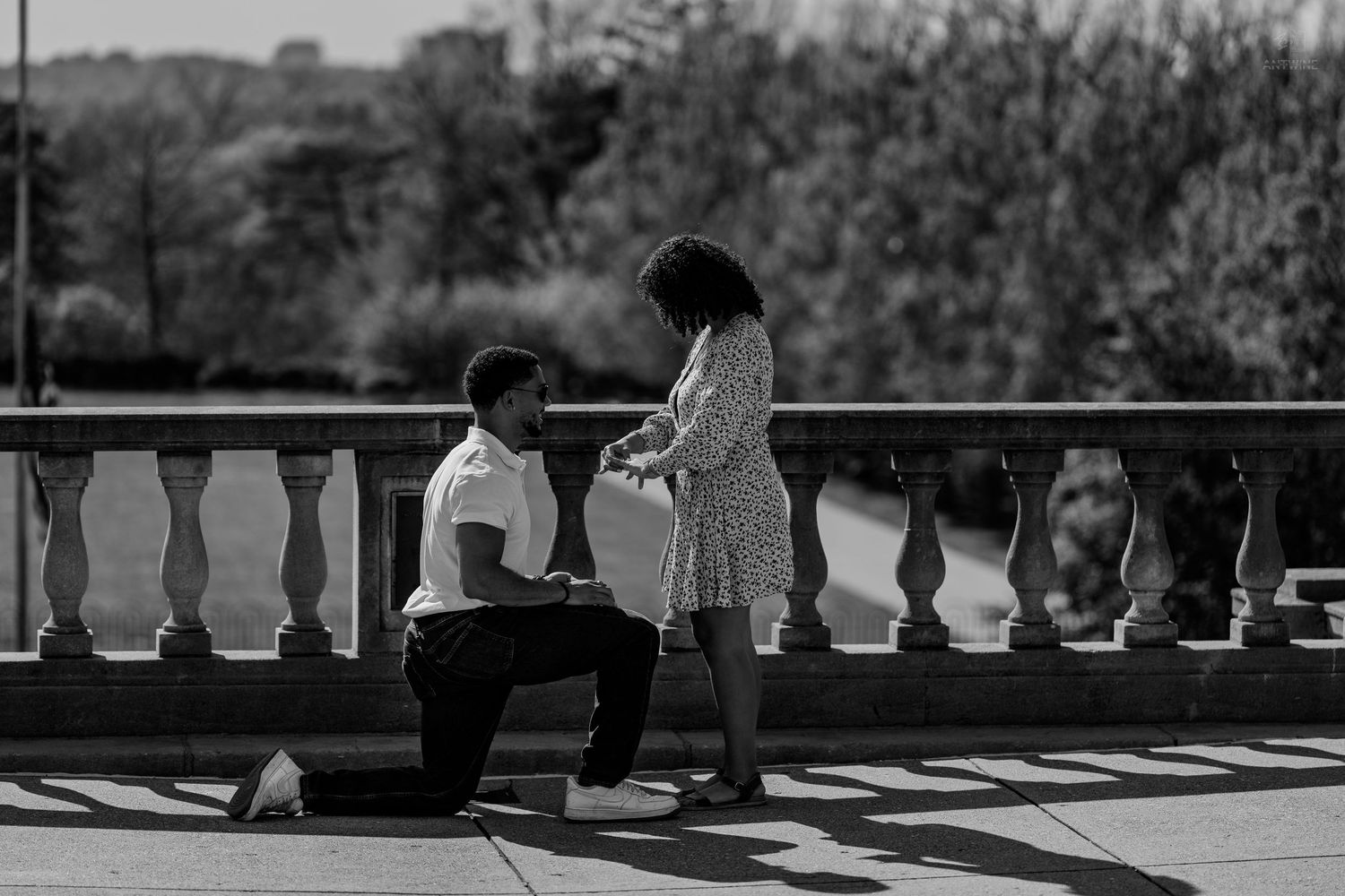 Black and white image of a man proposing on one knee to a woman on an outdoor stone terrace.