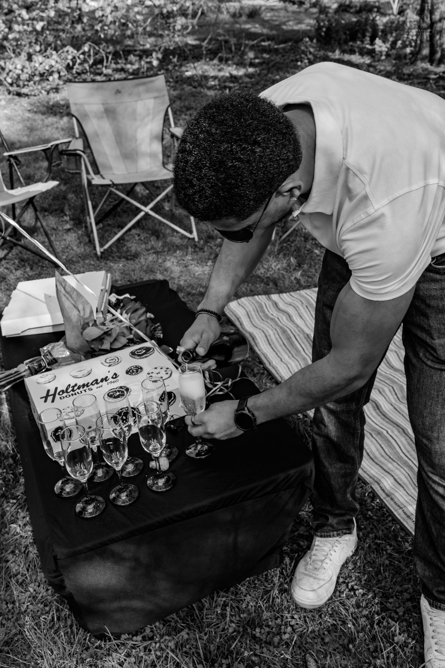 Man in white t-shirt arranging champagne flutes on a black table at an outdoor celebration.